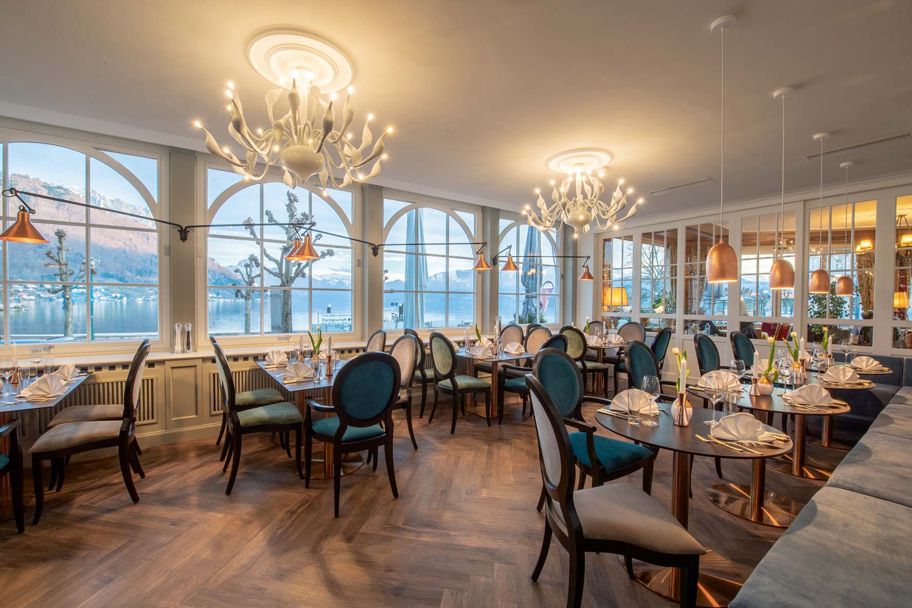 Elegantes Restaurant mit Holzboden, blauen und grünen Stühlen, dekorativen Kronleuchtern und großen Fenstern mit Blick auf einen See und Berge.