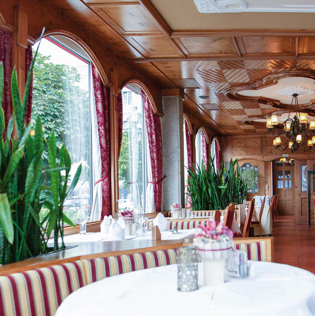 Elegantes Restaurant mit gestreiften Polstersitzen, weißen Tischdecken, großen Fenstern mit roten Vorhängen und vielen Grünpflanzen.