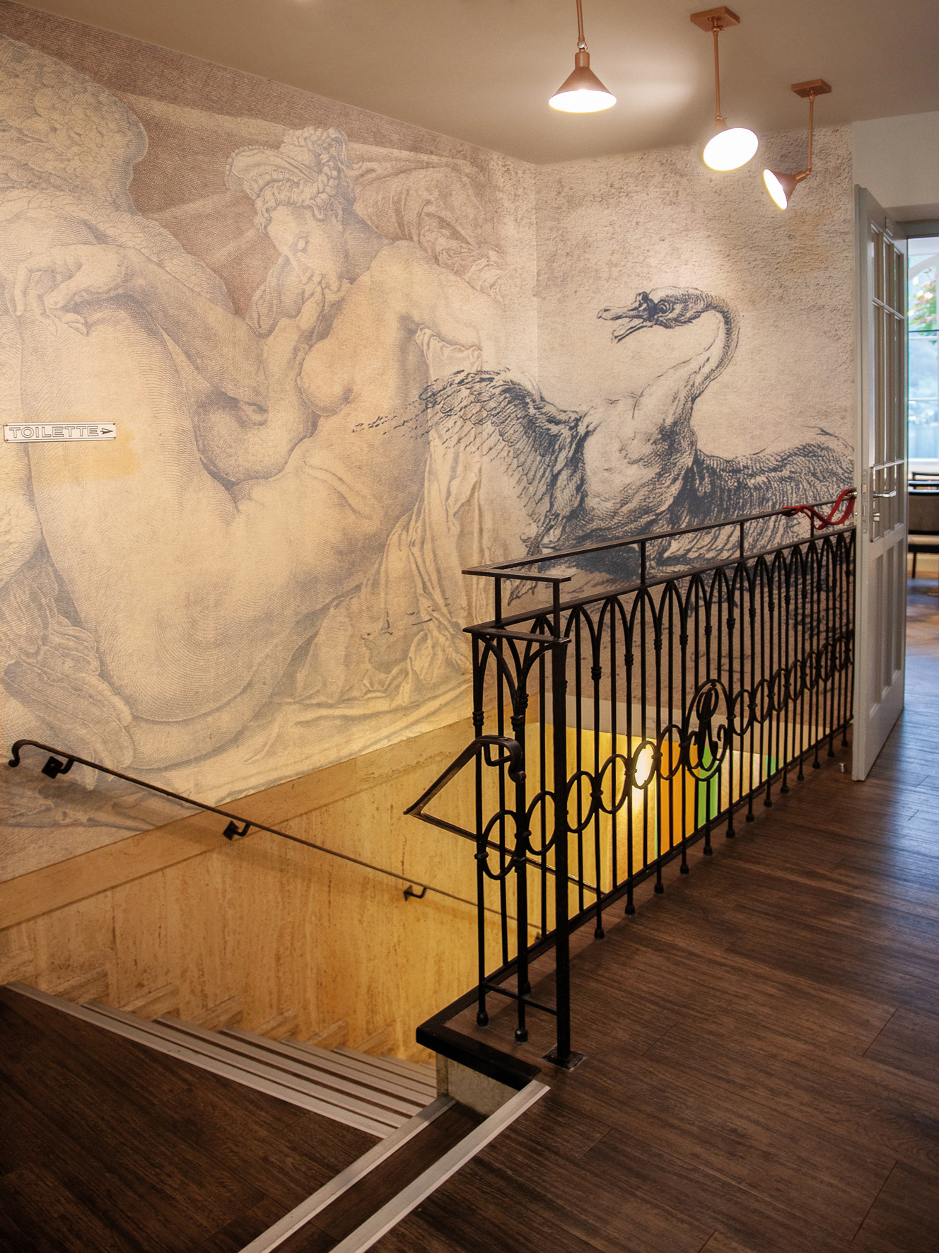 Treppenhaus mit einer Wandmalerei einer liegenden nackten Frau und einem ausgebreiteten Schwan, daneben ein Schild mit der Aufschrift 'Toilette'.