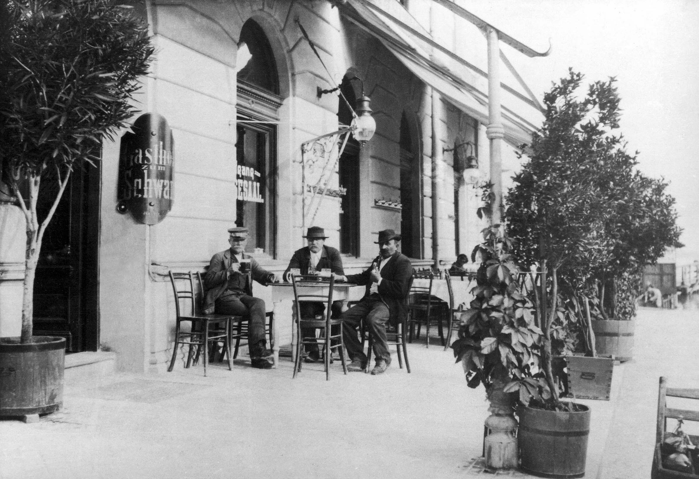 Drei Männer sitzen an einem Tisch vor einem Gastgarten um 1900, umgeben von Pflanzen in Kübeln.
