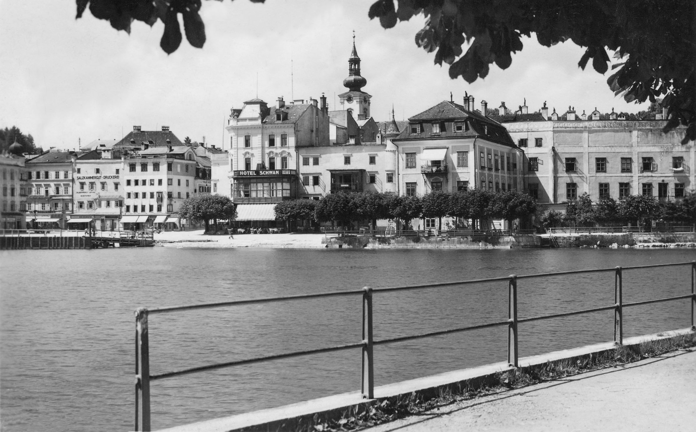 Schwarz-Weiß-Foto der Uferpromenade mit historischen Gebäuden, darunter das Hotel Schwan, und einem Kirchturm im Hintergrund.