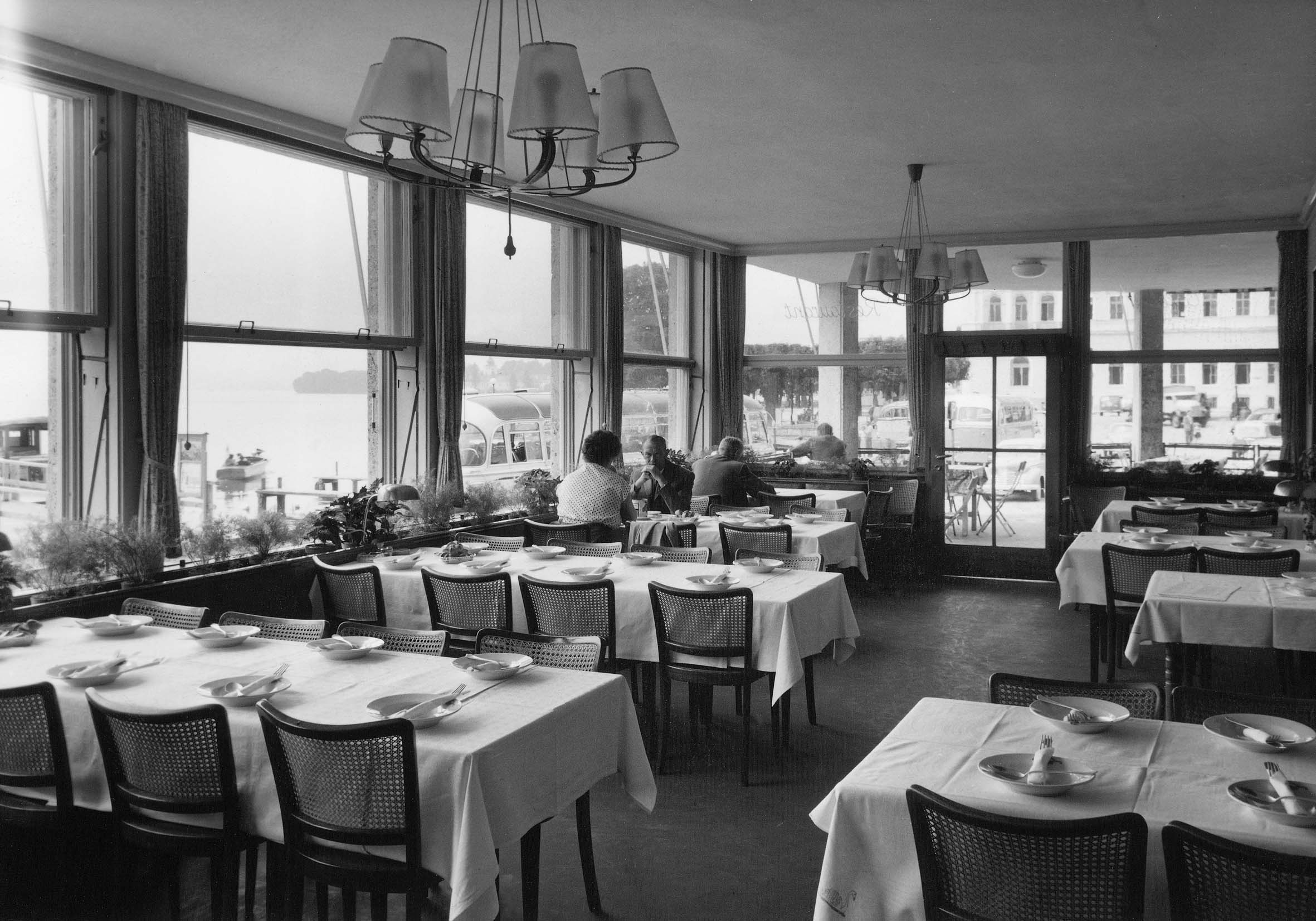 Schwarzweißfoto eines Speisezimmers mit gedeckten Tischen und Menschen, die durch große Fenster auf einen Hafen und parkende Autos blicken.