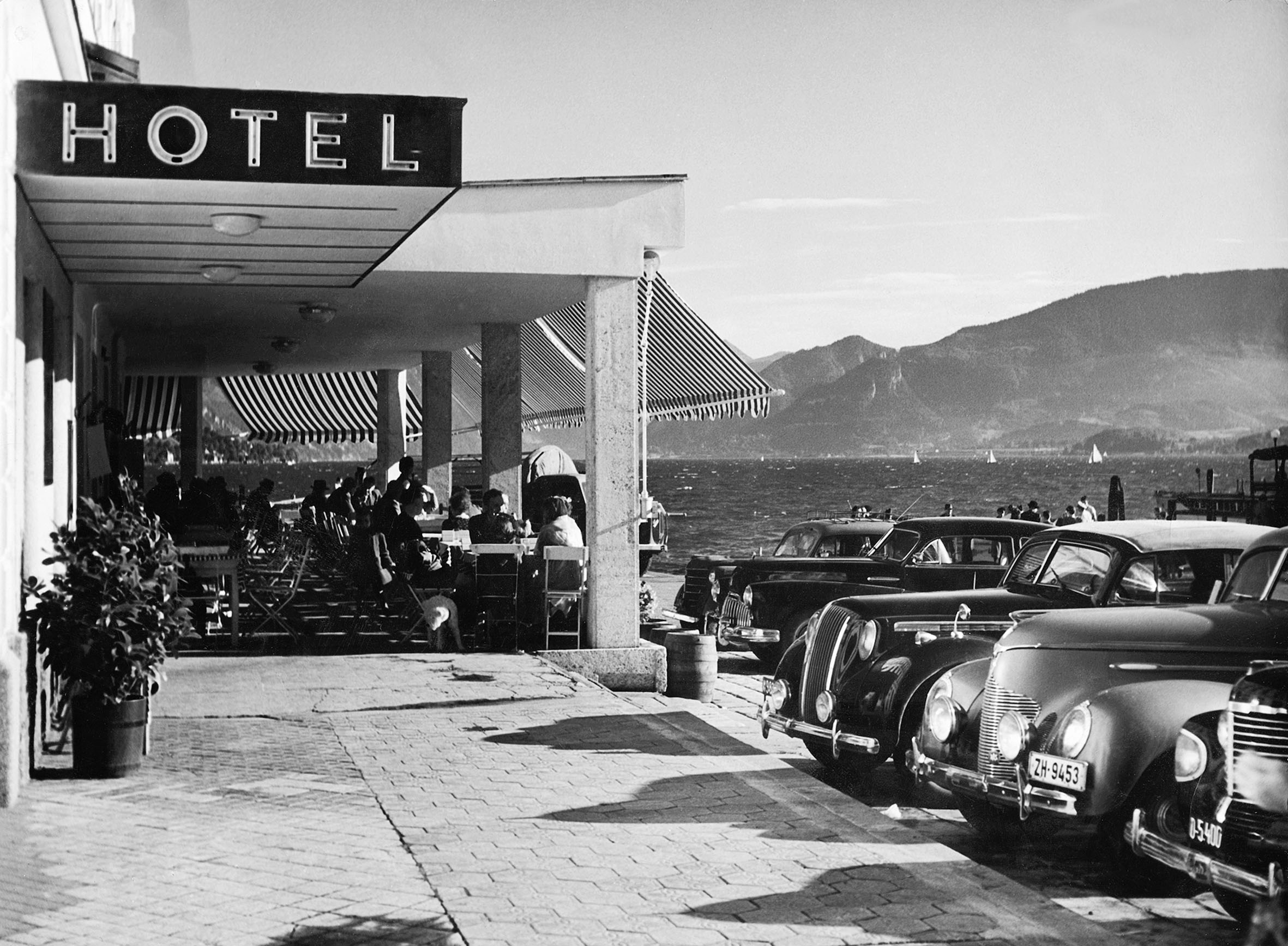 Schwarz-weiß-Foto eines Hotels mit Gästen auf der Terrasse und geparkten klassischen Autos neben einem See mit Bergen im Hintergrund.