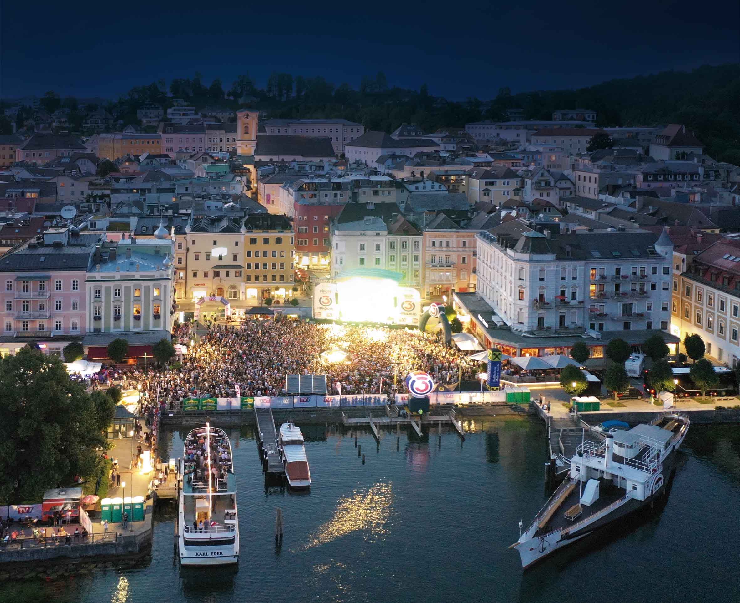 Nächtliches Stadtbild von Gmunden mit einem großen, beleuchteten Konzertpublikum am Seeufer und zwei Schiffen im Wasser.