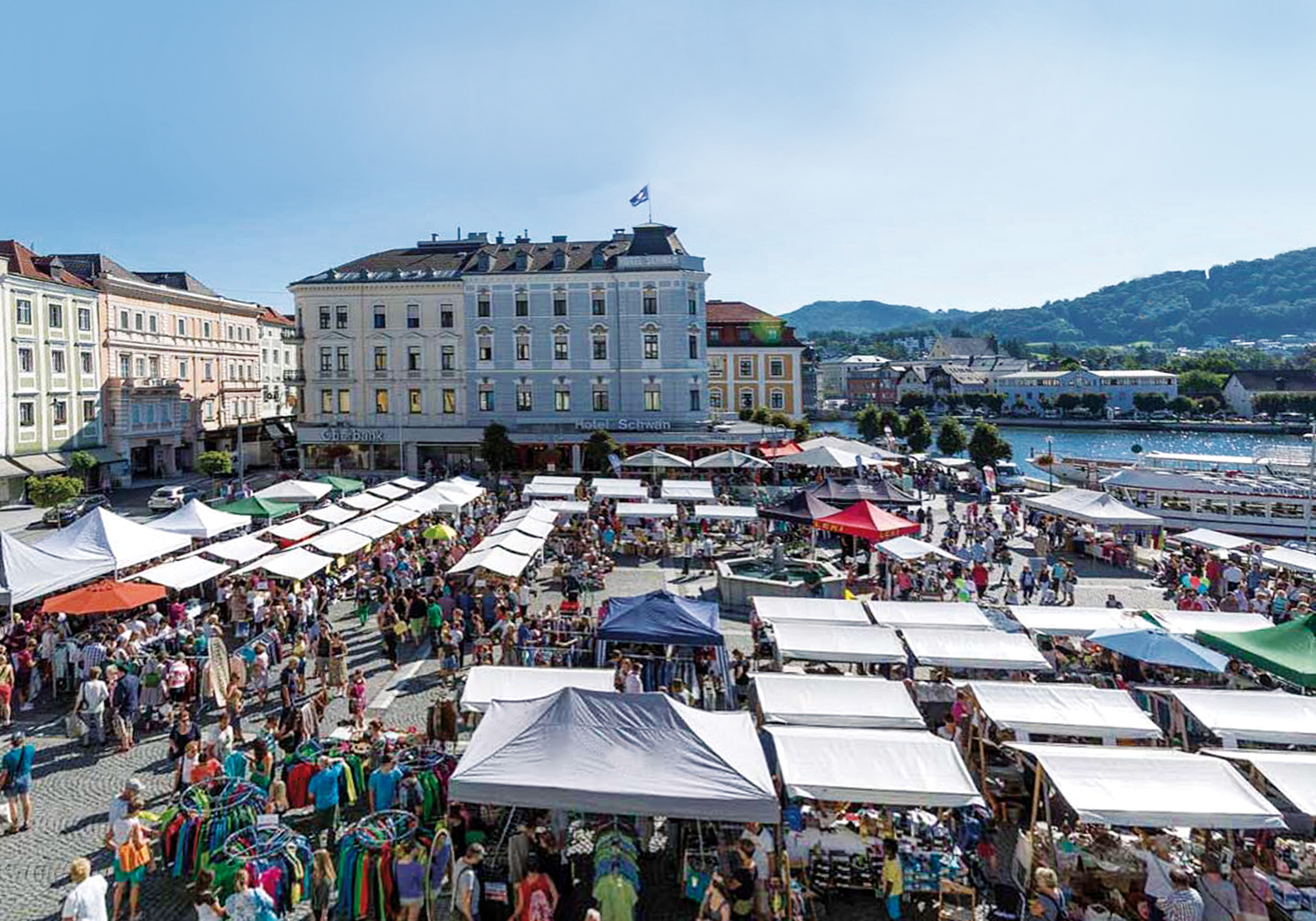 Belebter Wochenmarkt mit vielen Ständen und Menschen vor historischen Gebäuden nahe einem Fluss.
