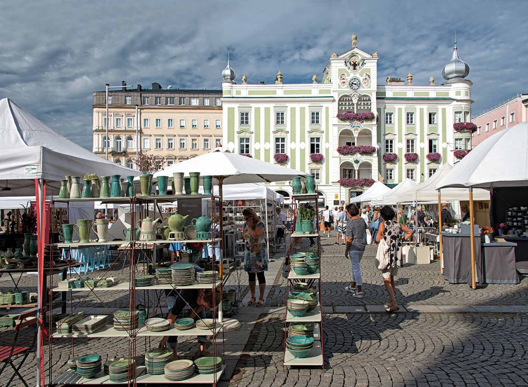 Marktstände mit Keramikgeschirr auf Pflasterplatz vor historischer, mit Blumen geschmückter Fassade unter bewölktem Himmel.