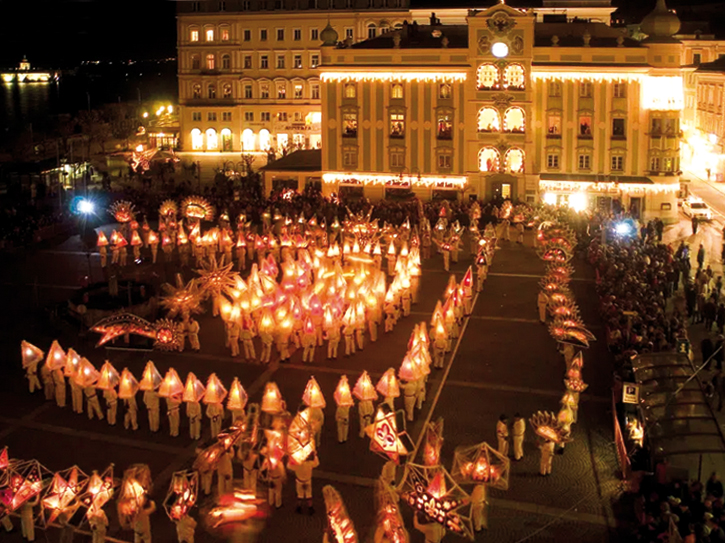 Nächtliche Prozession mit zahlreichen Menschen in weißen Gewändern, die erleuchtete Laternen in Form von Glocken tragen, vor einem historischen, beleuchteten Gebäude.