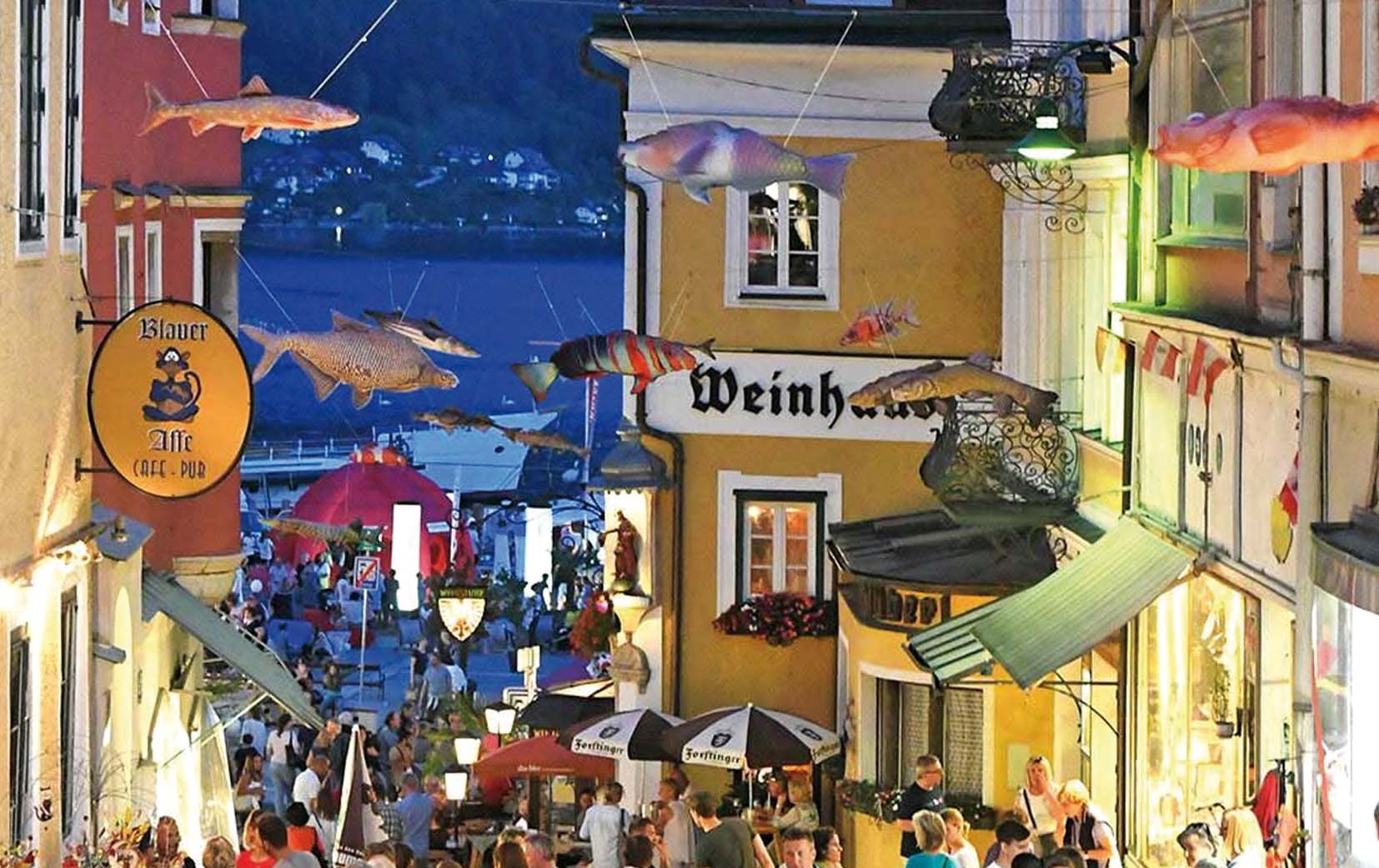 Belebte Gasse mit Menschen, bunten Gebäuden und hängenden Fischen als Dekoration bei Abenddämmerung am Seeufer.