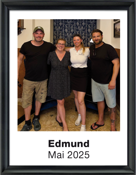 Vier lächelnde Erwachsene, zwei Männer und zwei Frauen, posieren zusammen drinnen vor einem Wandteppich. Darunter steht der Name Edmund und Mai 2025.