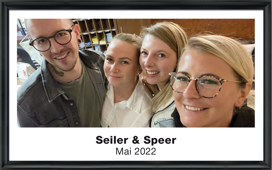 Vier lächelnde Personen, zwei Frauen und zwei Männer, stehen eng zusammen für ein Foto in einem Raum mit einem Regal im Hintergrund.