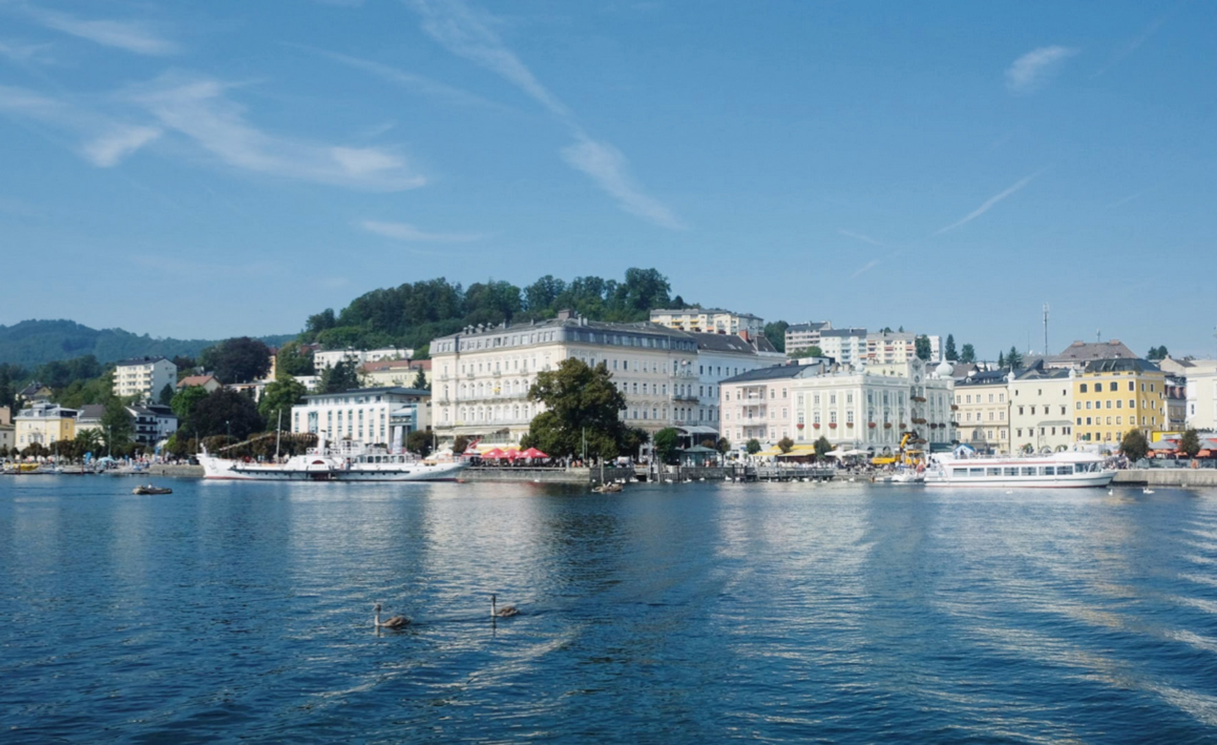 Blick auf den Uferbereich einer Stadt mit weißen und gelben Gebäuden, mehreren Booten im Wasser und Bergen im Hintergrund unter blauem Himmel.