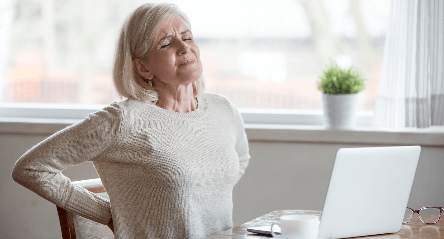 Senior woman experiencing lower back pain while sitting at a table, illustrating symptoms that may require disc replacement surgery and care from the best spine surgeon in Los Angeles.