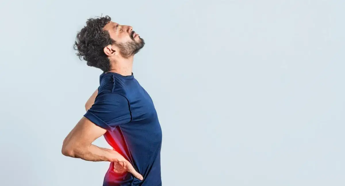 Adult standing with a hand on the lower back, suggesting lumbar strain or lower back pain