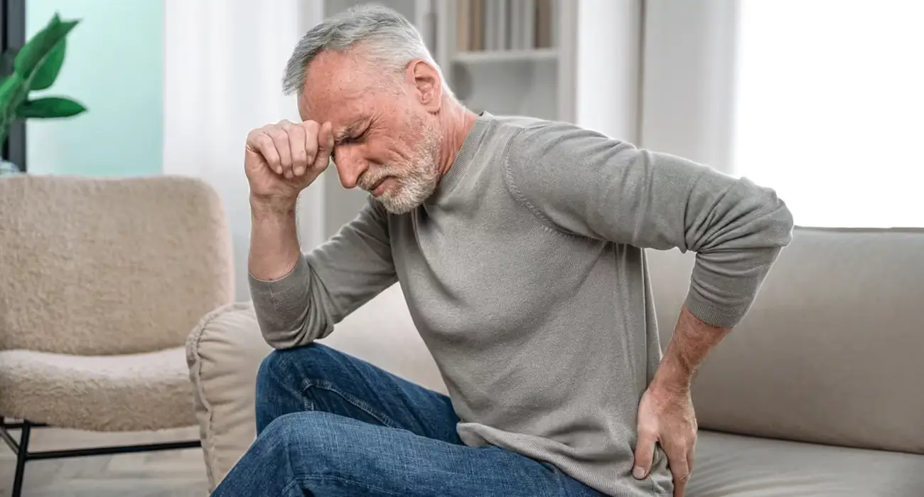 Senior man sitting on a couch holding his lower back in pain, illustrating lower back pain treated by the best spine surgeon for artificial disc replacement in Los Angeles