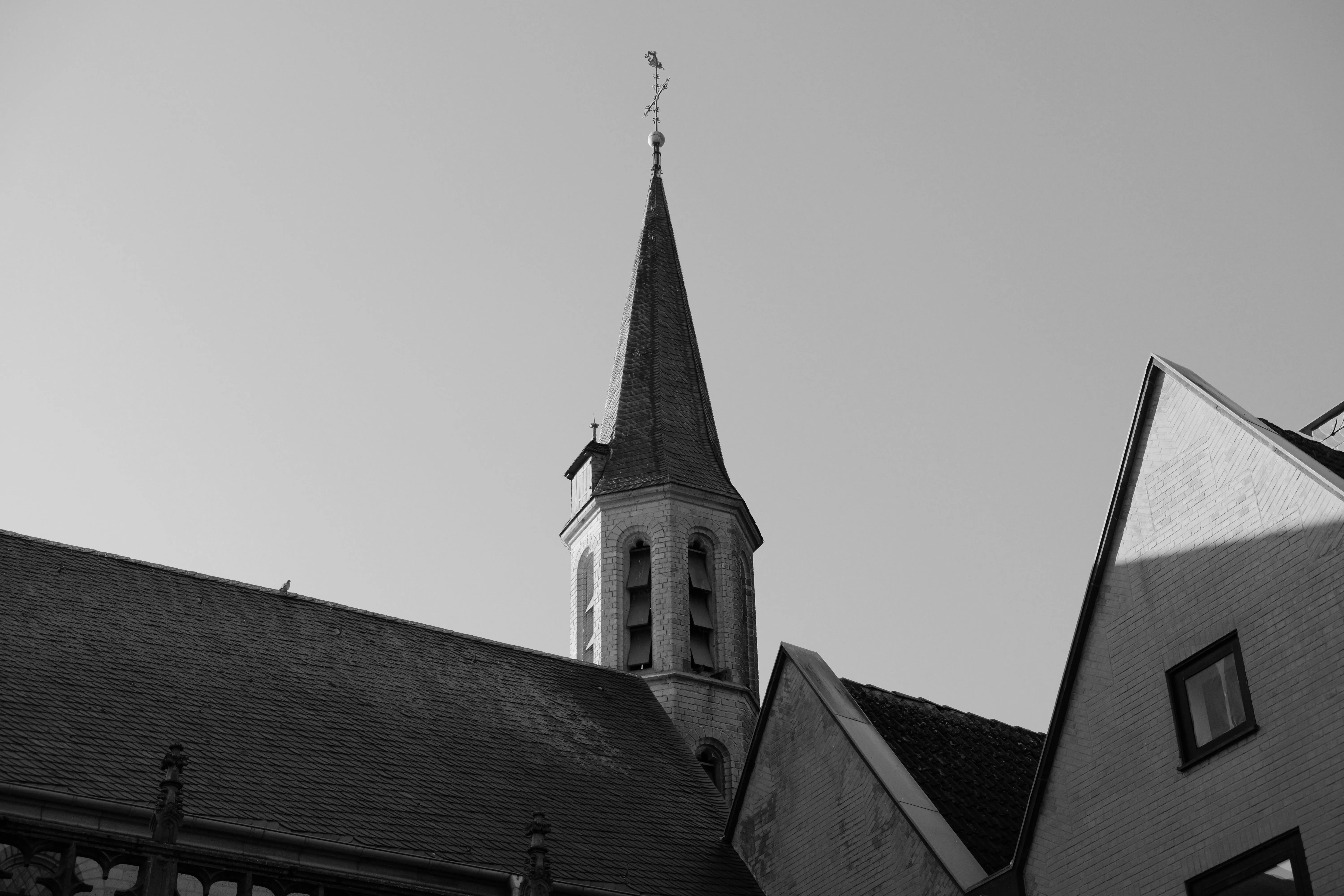 Black and white photo of a church steeple with a weather vane, surrounded by rooftops under a clear sky.