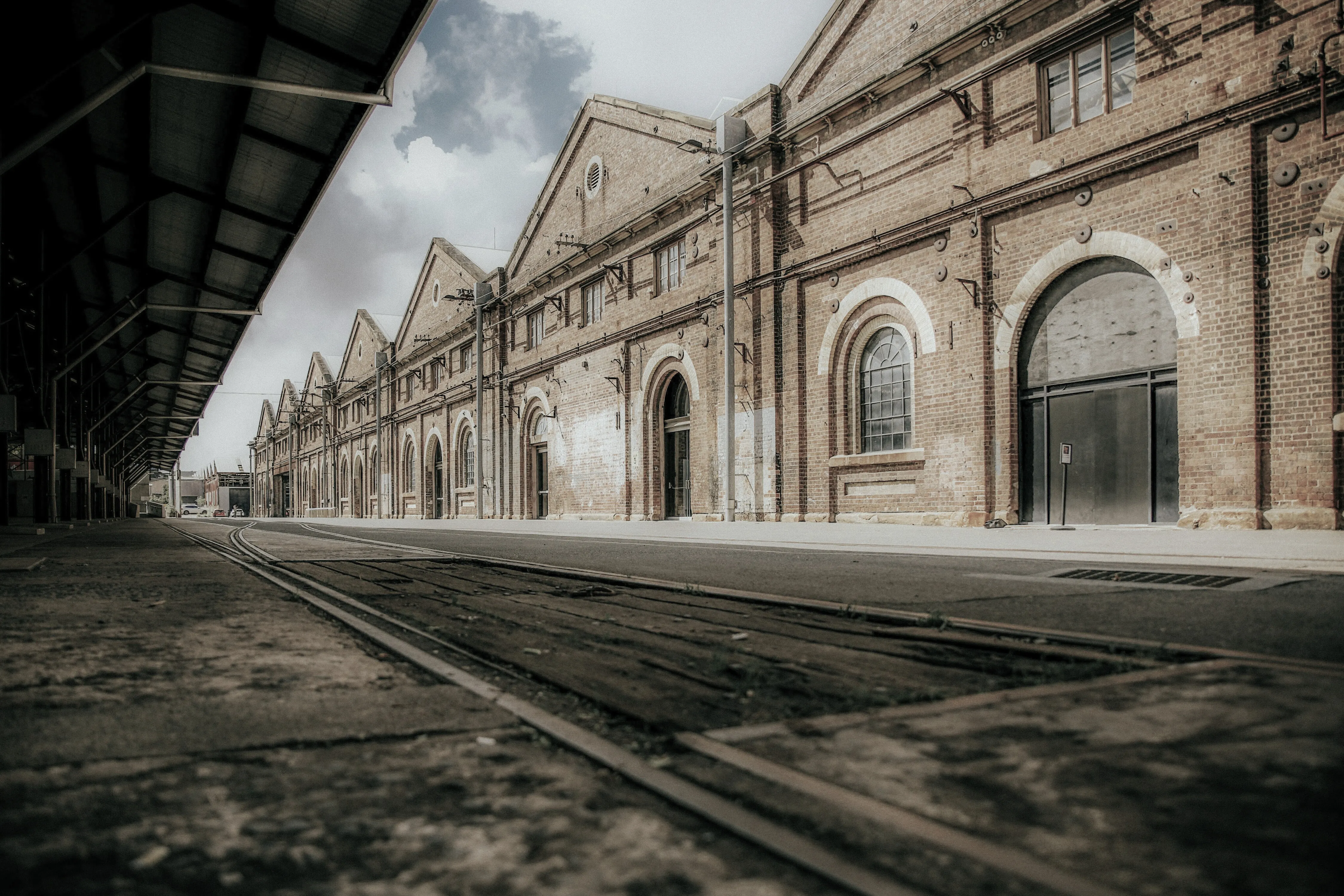 Long row of old brick warehouse buildings with arched windows and doors along empty street with train tracks.