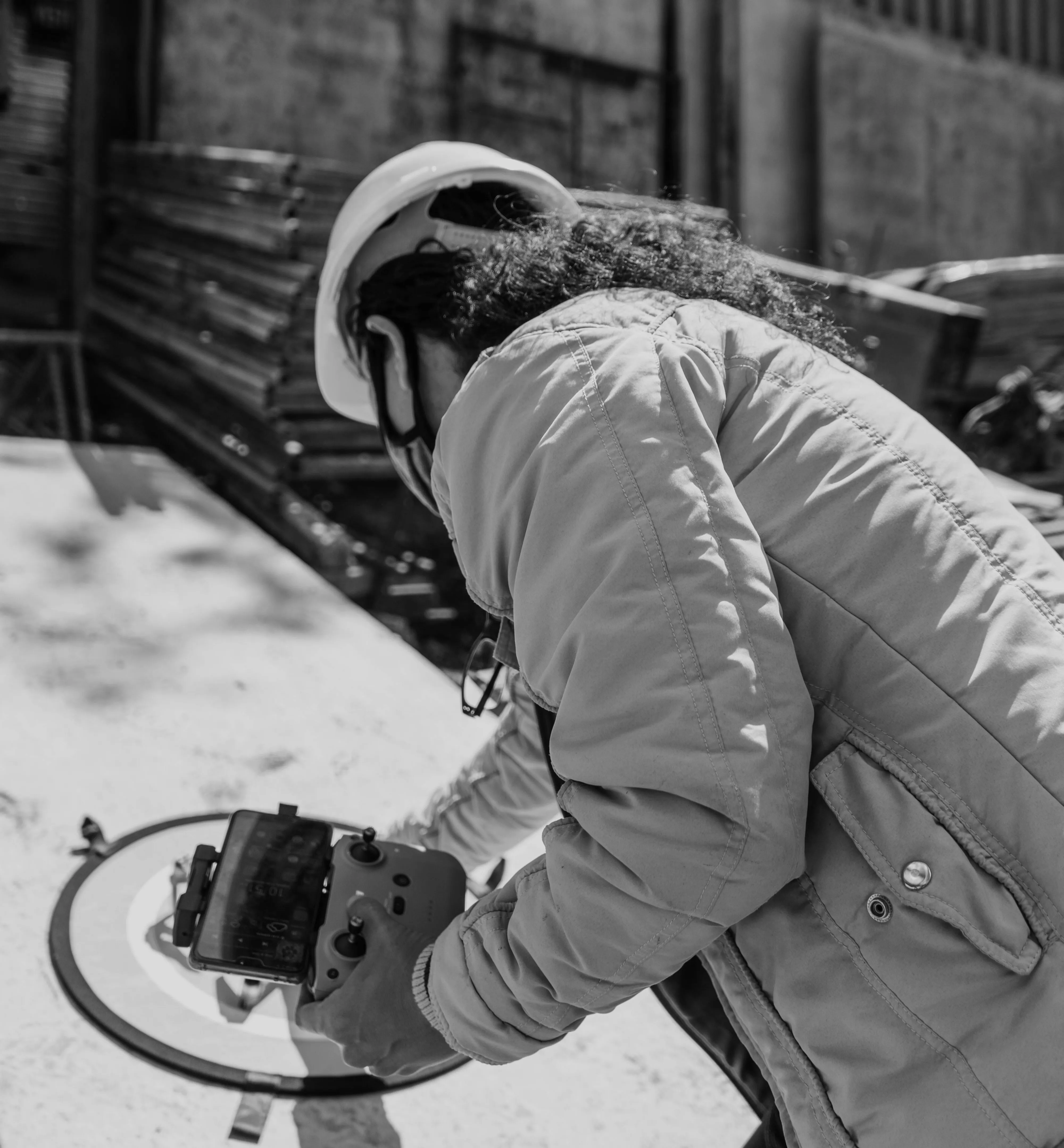 Person in a jacket and helmet operating a remote controller with a screen next to a drone landing pad.
