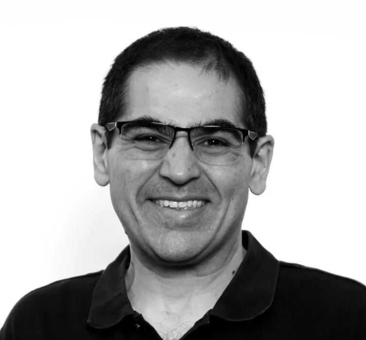 Smiling man with short dark hair, glasses, and a dark collared shirt against a plain white background.