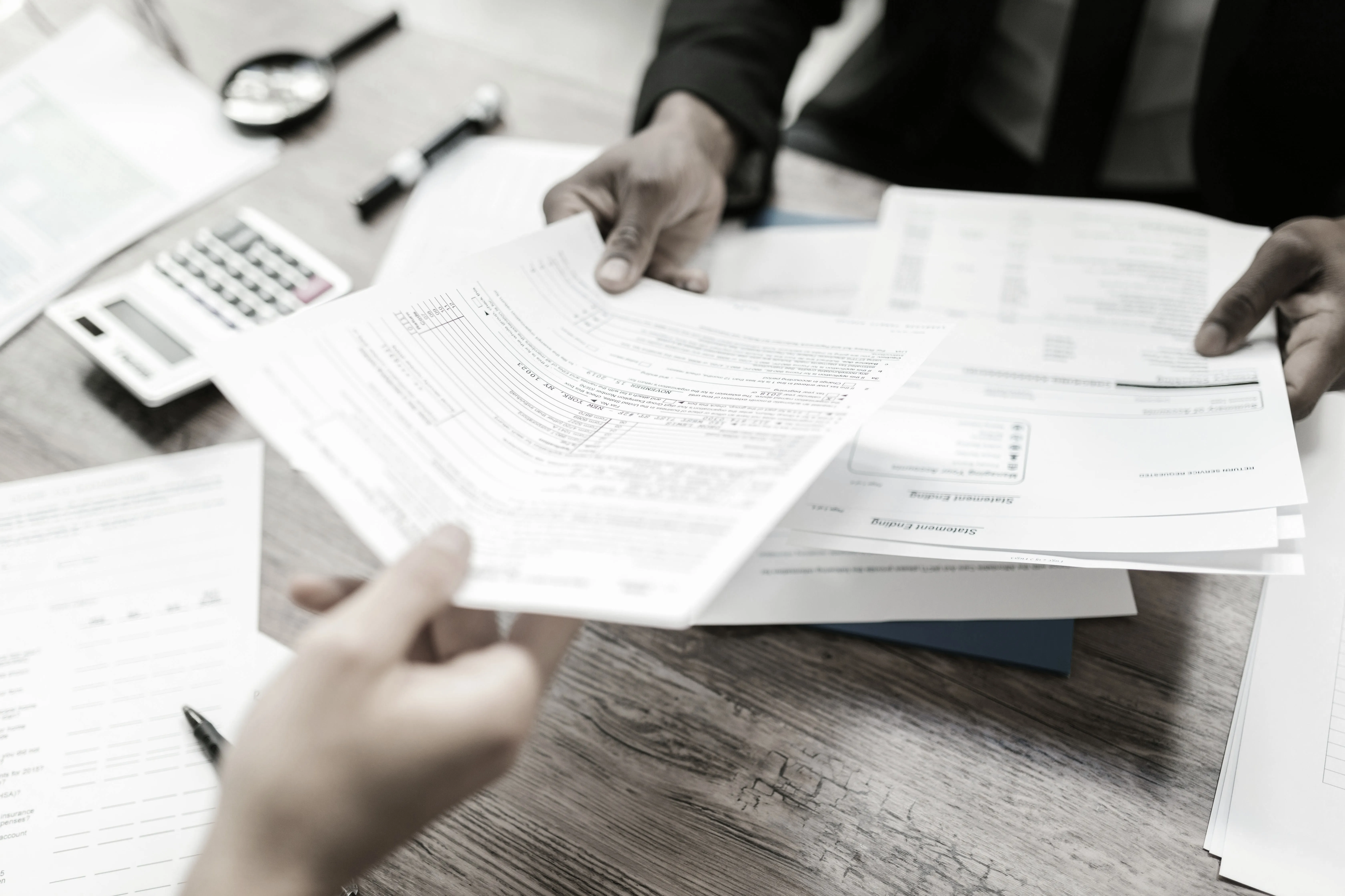 Two people exchanging financial documents over a wooden table with a calculator and pen nearby.