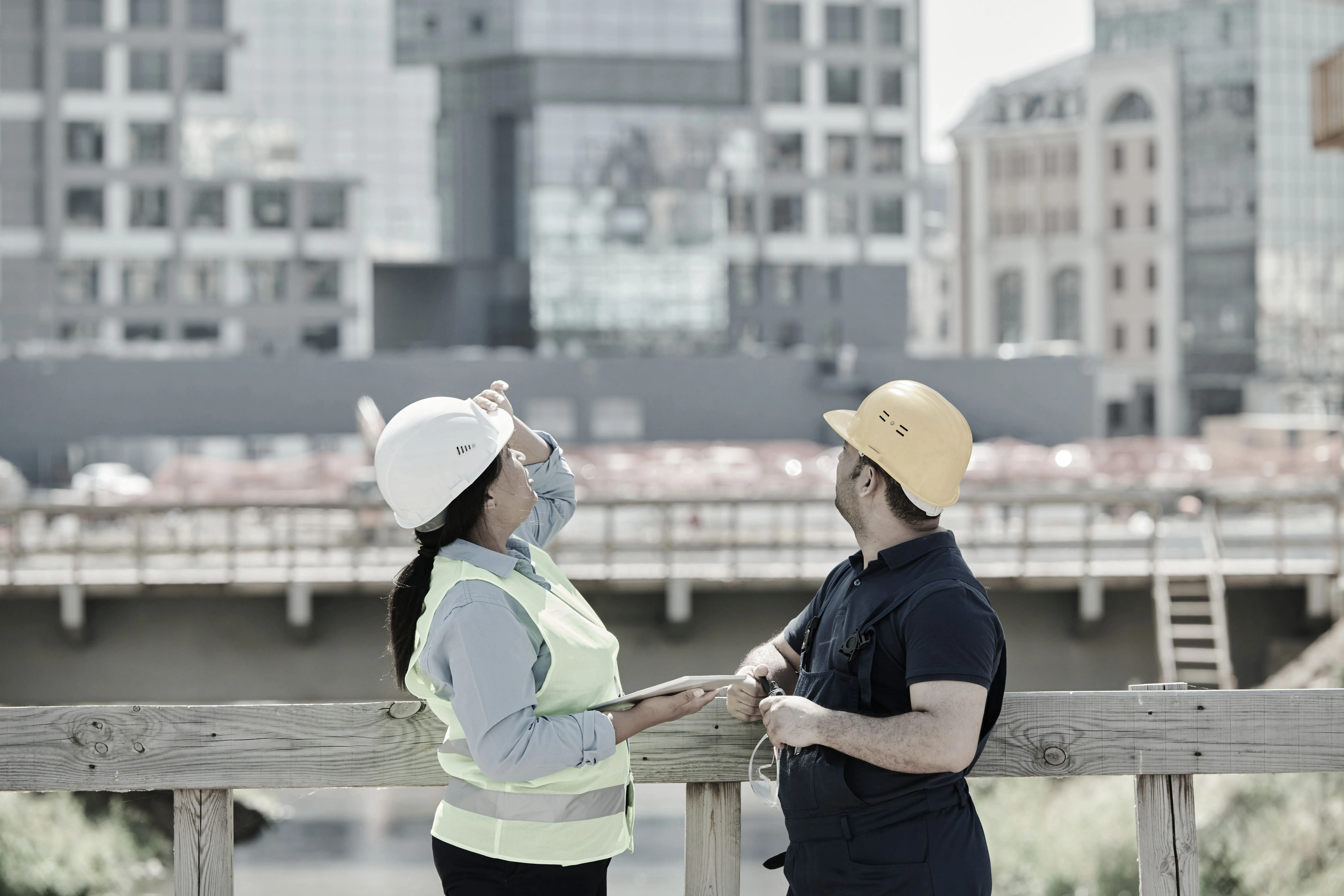 Two construction workers wearing helmets and safety vests discussing project plans at a site with buildings in the background.