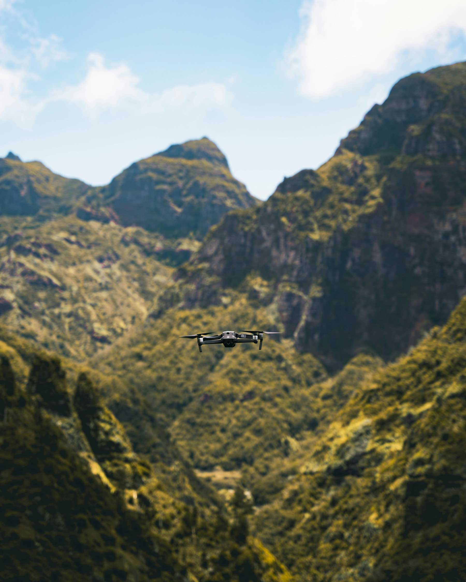 Drone flying in front of a green mountainous landscape under a partly cloudy blue sky.