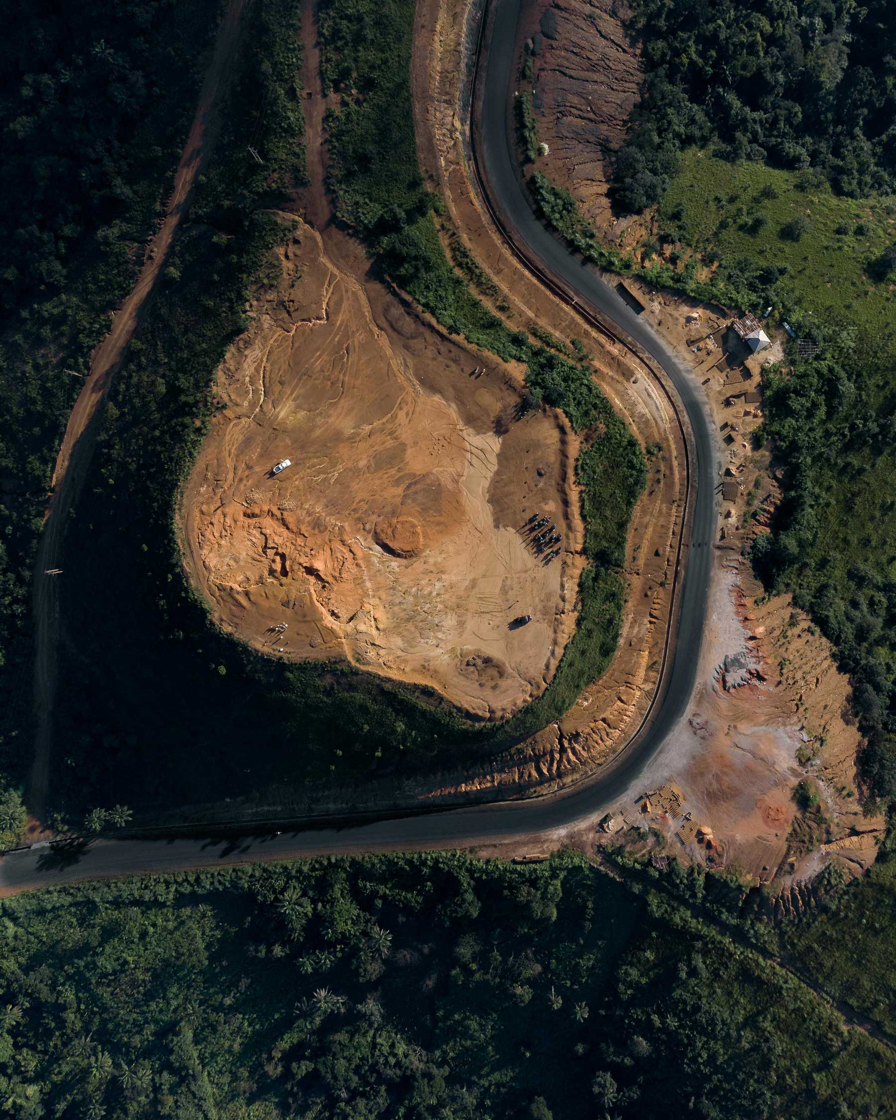 Aerial view of a winding road curving around a cleared dirt area surrounded by dense green forest.