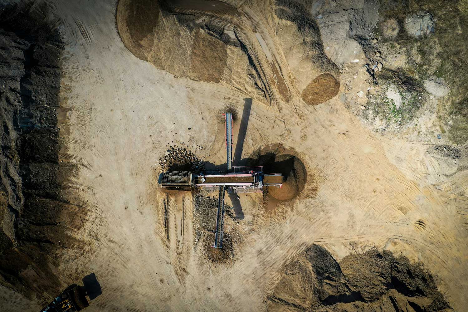 Aerial view of a dirt construction site with a large piece of machinery sorting soil and gravel into separate piles.