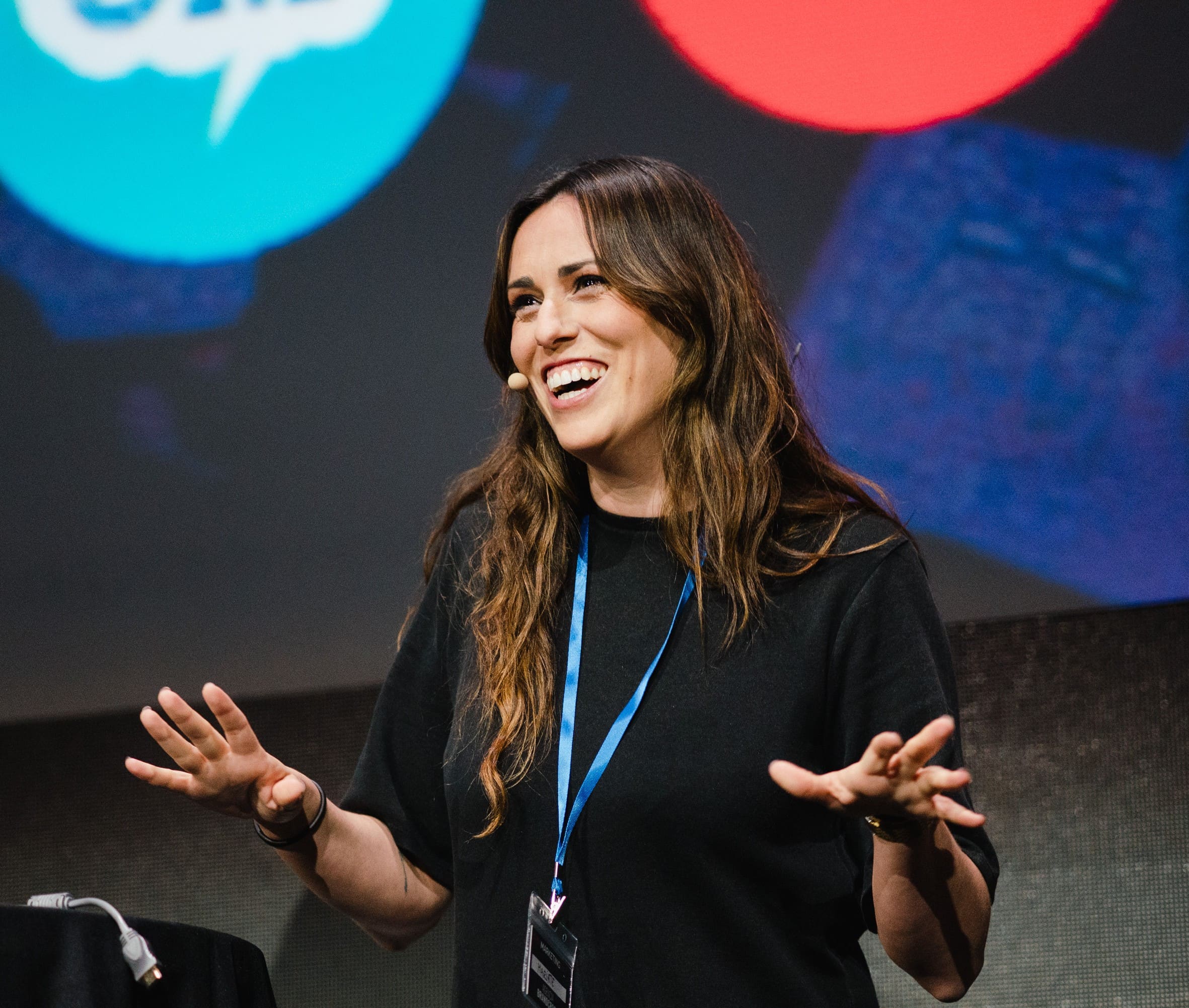 Smiling woman with long brown hair speaking on stage wearing a black shirt and blue lanyard.