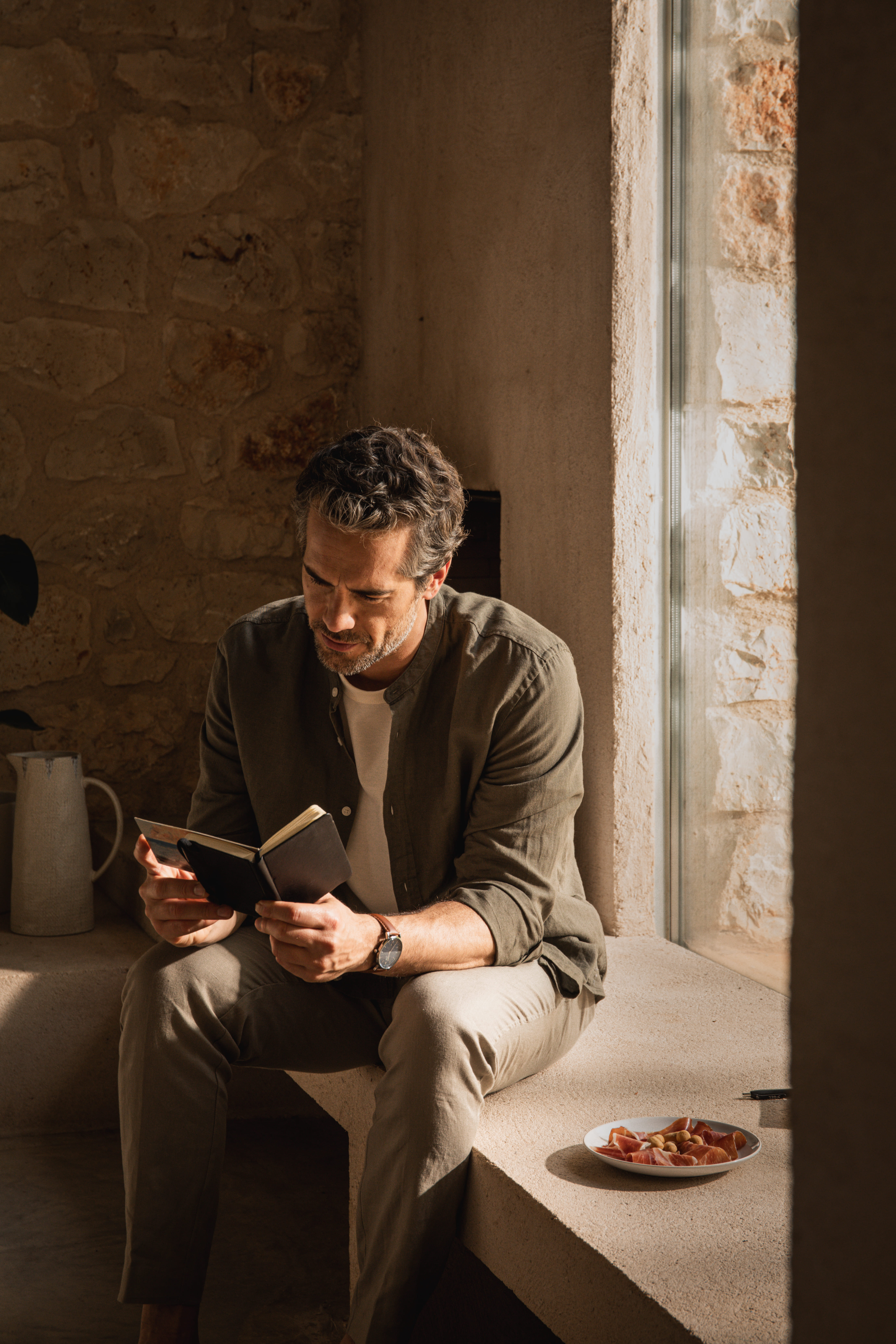 Man sitting by a sunlit stone window ledge reading a small book, with a plate of sliced meat beside him.