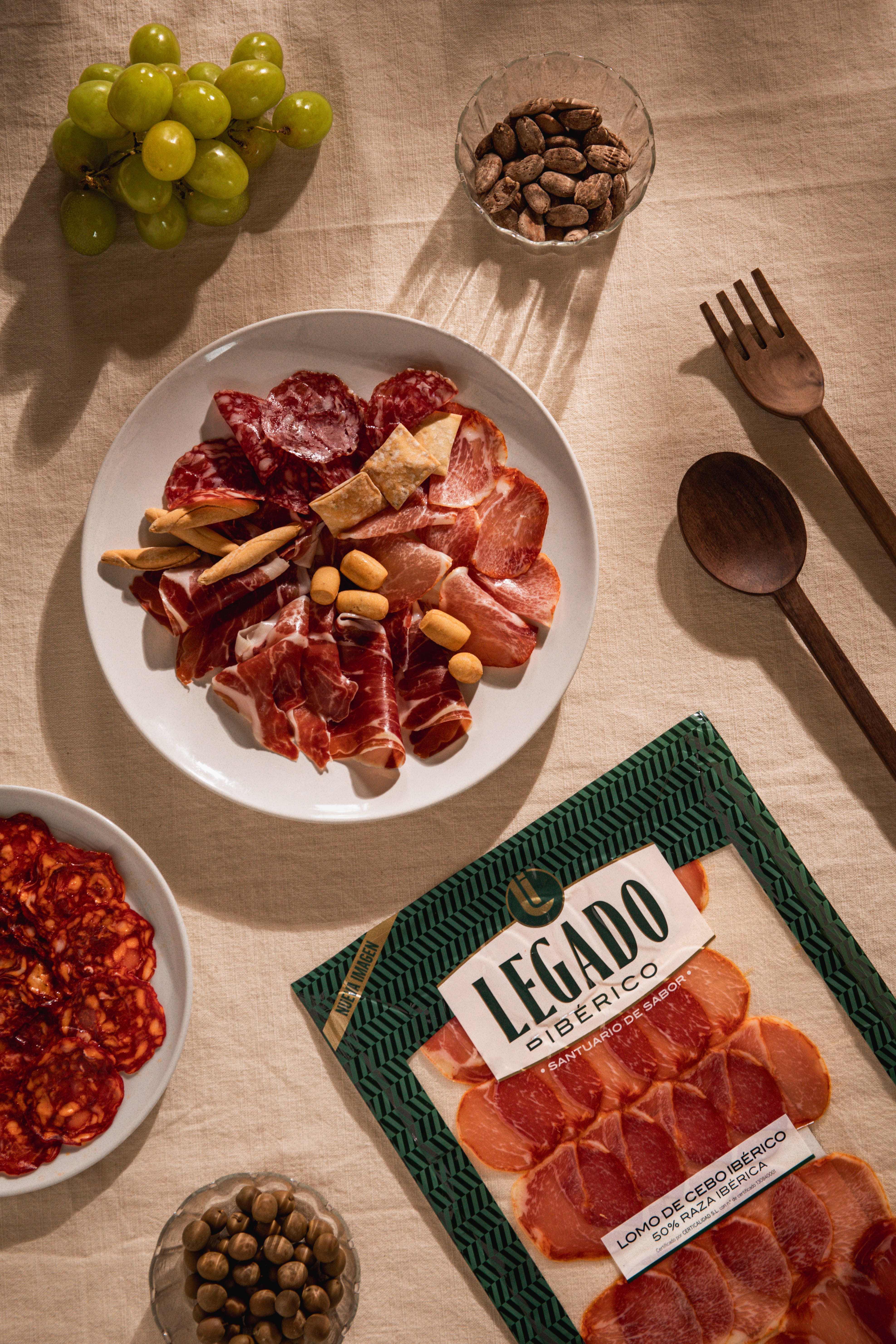 Flat lay of sliced cured meats on a white plate and in packaging, with green grapes, olives, nuts in a glass bowl, and wooden utensils on a beige tablecloth.