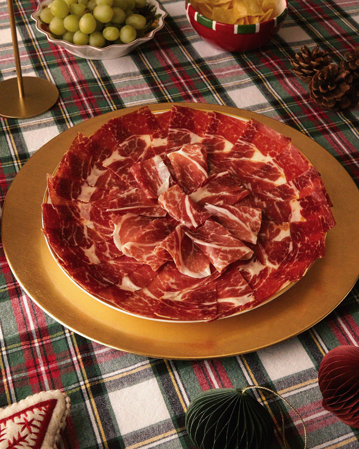 Plate of thinly sliced cured meat arranged in a circular pattern on a gold charger, on a plaid tablecloth with bowls of green grapes and chips nearby.