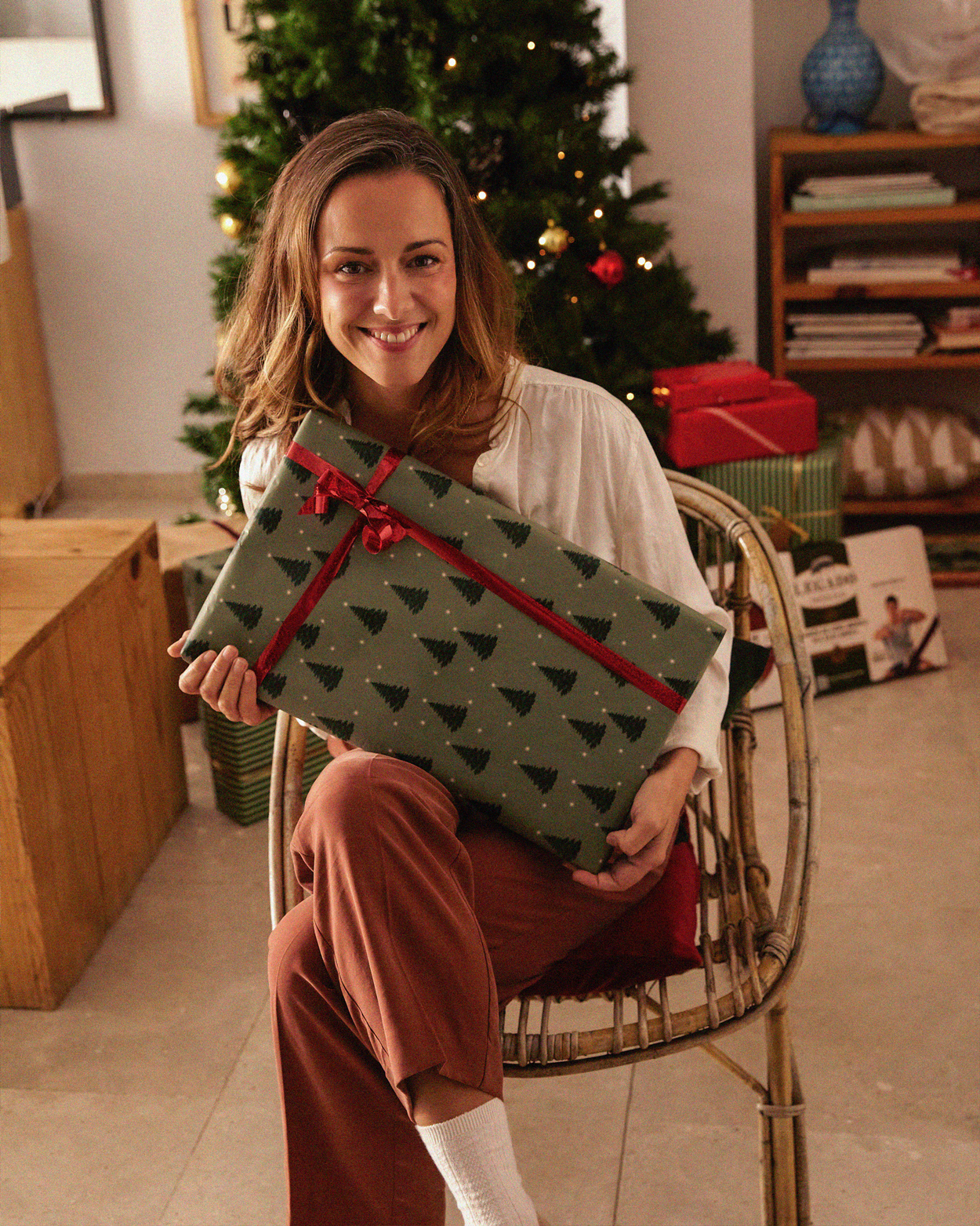 Smiling woman sitting on a chair holding a wrapped Christmas gift with a decorated Christmas tree in the background.