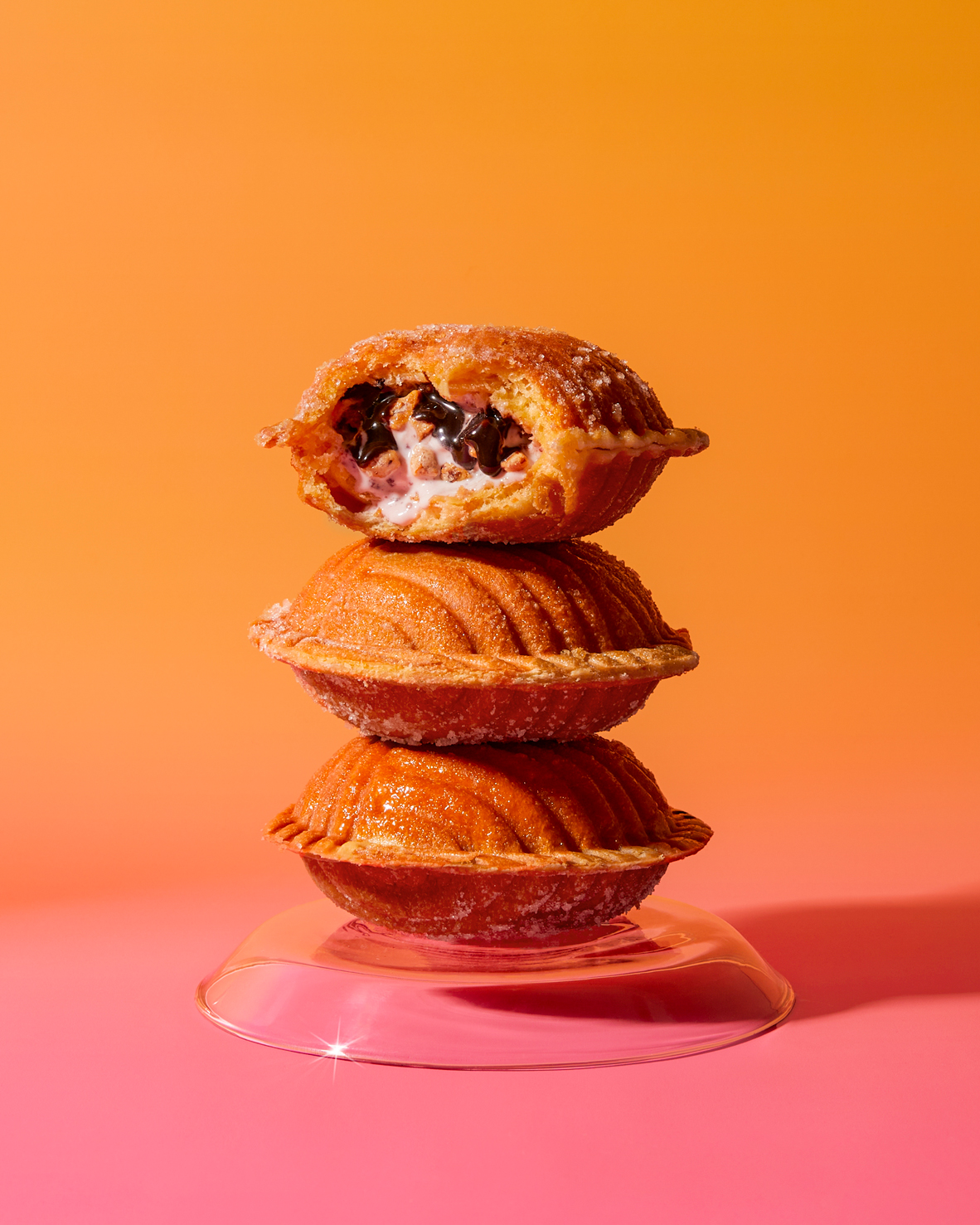 Stack of three sugar-coated pastries with one cut open to reveal creamy chocolate and pink filling, displayed on a clear stand against an orange and pink gradient background.