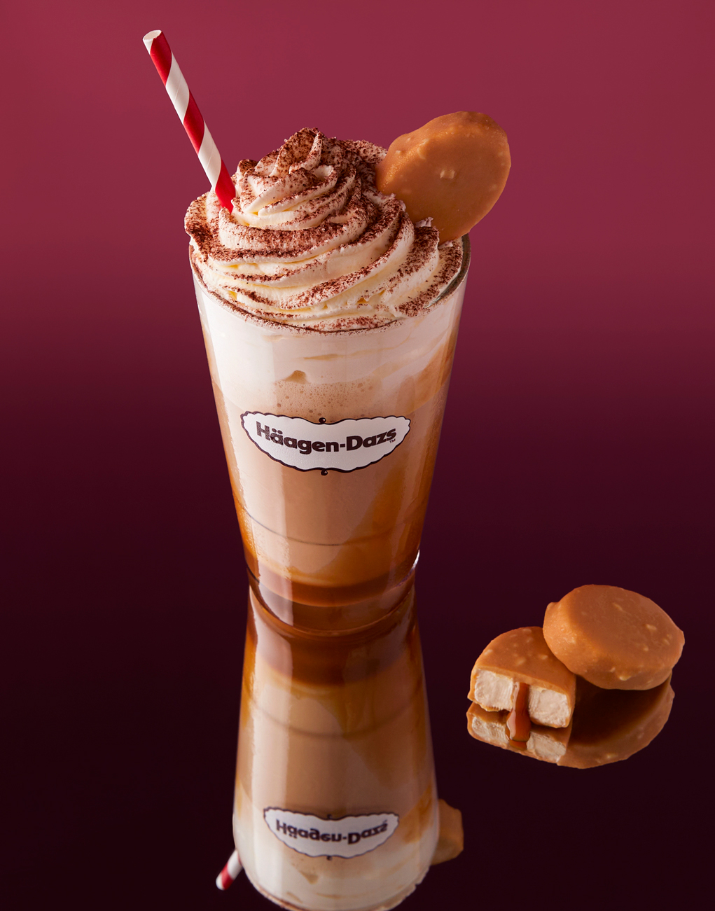 Glass of Häagen-Dazs caramel drink topped with whipped cream, cocoa powder, red and white striped straw, and a caramel-covered ice cream bite with two more bites beside it.