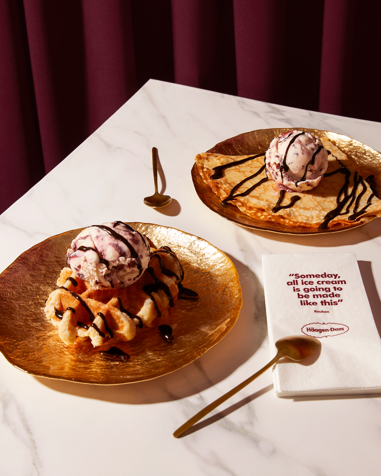 Two golden plates with desserts: one with a waffle topped with a scoop of ice cream drizzled with chocolate sauce, and one with a folded crepe topped with a scoop of ice cream and chocolate drizzle, set on a white marble table with gold spoons and a napkin displaying a Häagen-Dazs quote.