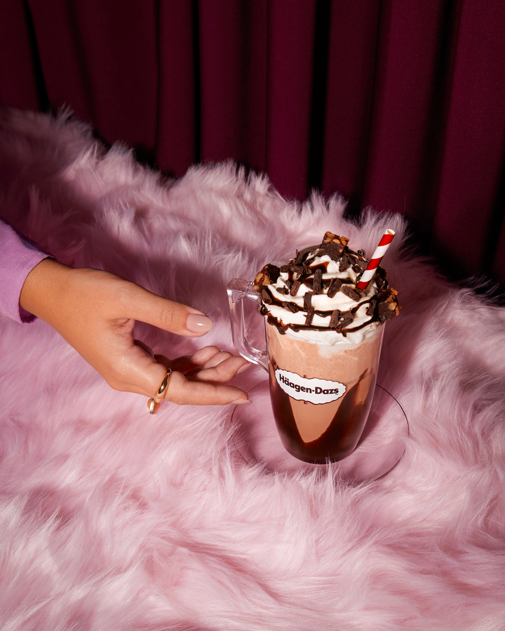 Hand presenting a glass cup of Häagen-Dazs hot chocolate topped with whipped cream, chocolate shavings, and pieces of brownies on a pink fluffy surface.