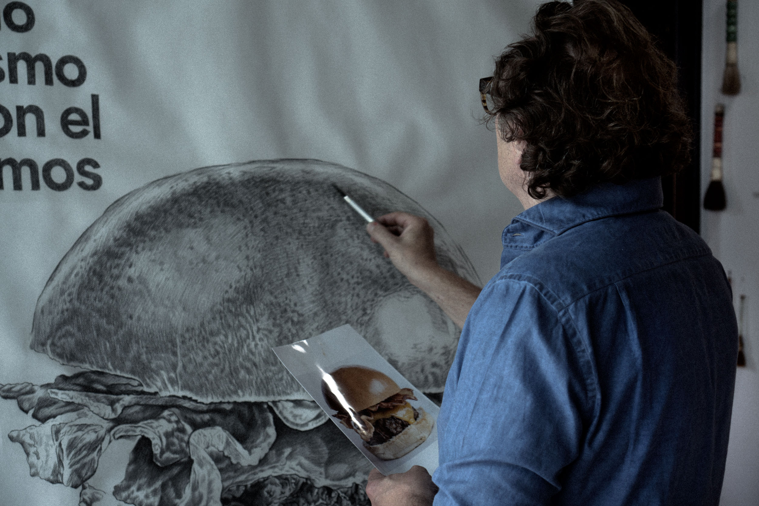 Person with curly hair in a blue shirt painting a large black-and-white burger illustration while holding a photo of a real burger.