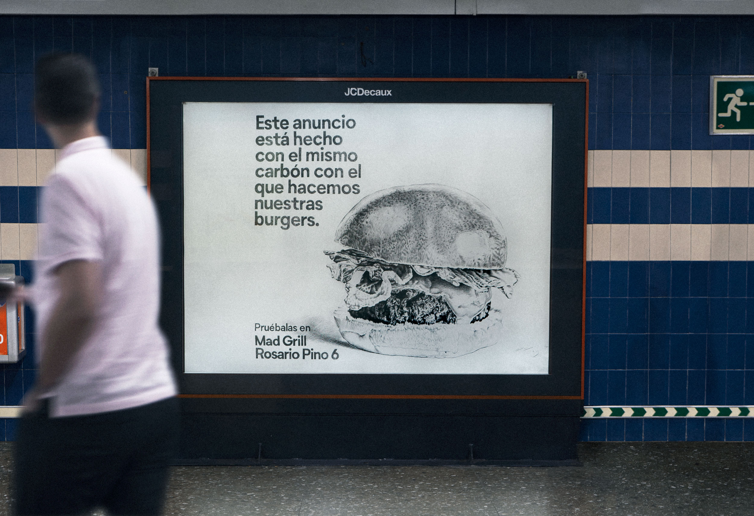 Advertisement in a subway station showing a detailed black and white sketch of a burger with text in Spanish promoting Mad Grill burgers made with the same charcoal used in the ad.