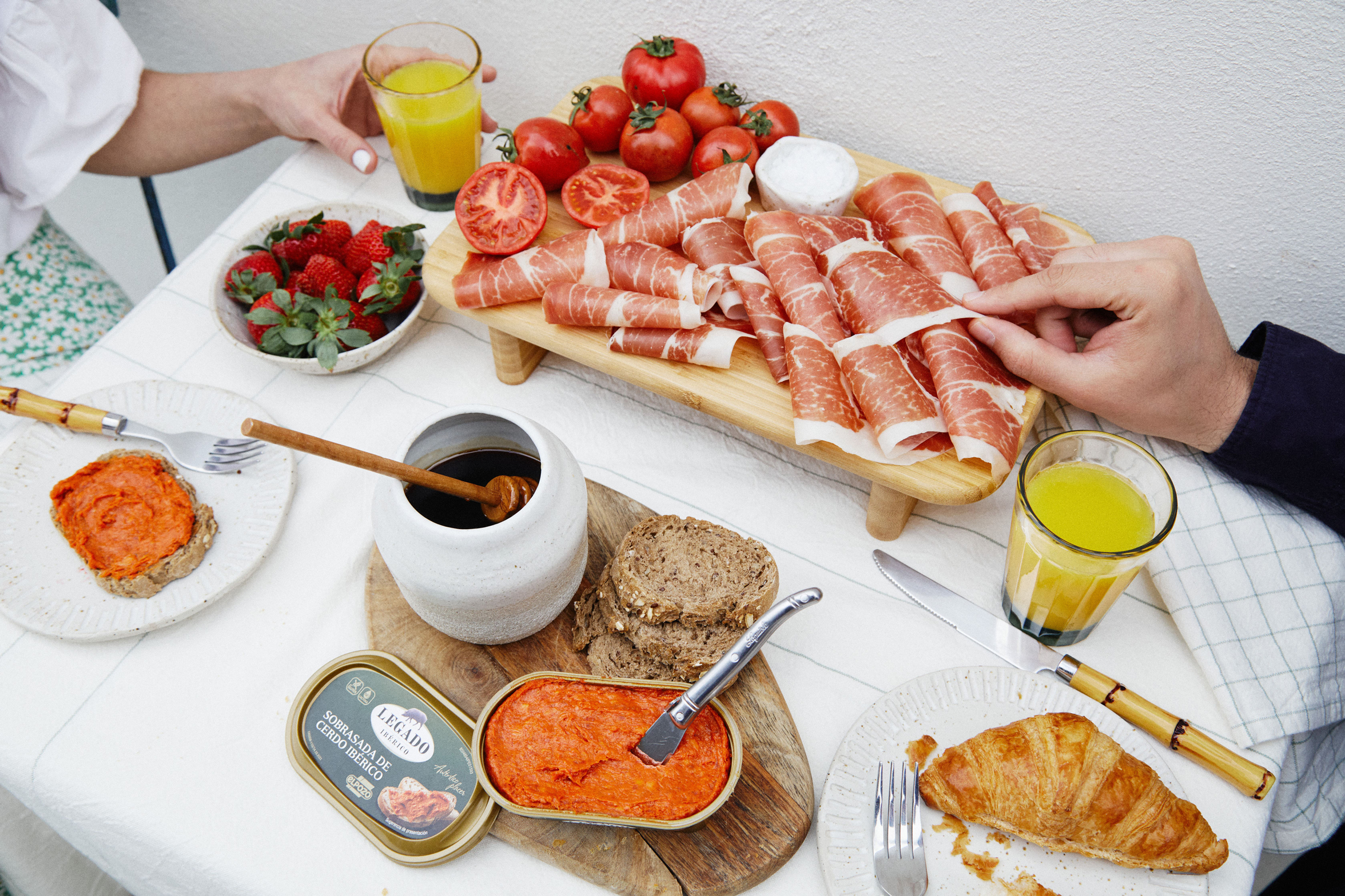 Table set with orange juice, bowl of strawberries, sliced tomatoes, rolled prosciutto, ospabad spread with bread, croissant on plates, and two people's hands.
