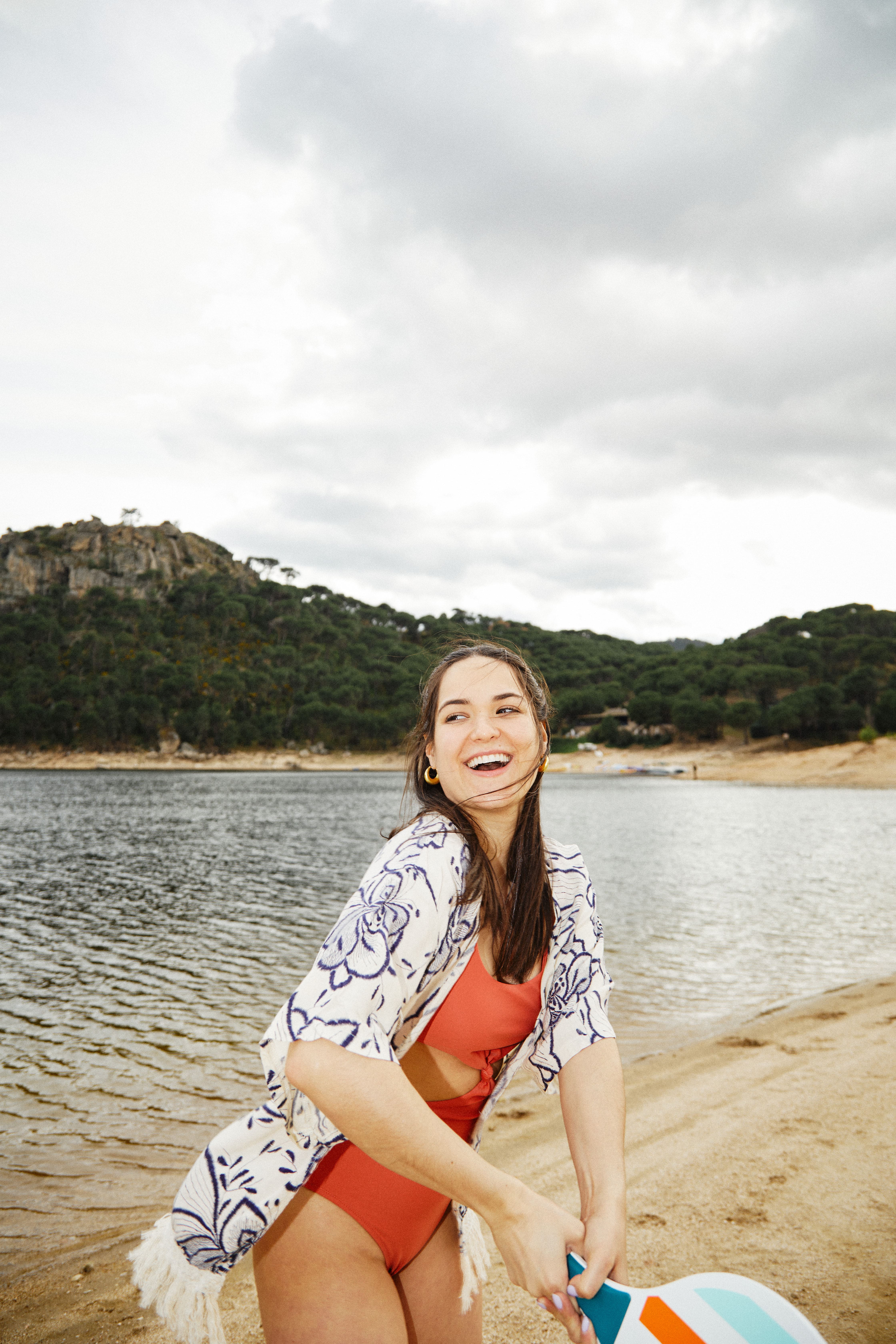 Smiling young woman in a red swimsuit and floral cover-up holding a paddle on a sandy lakeshore with hills in the background.