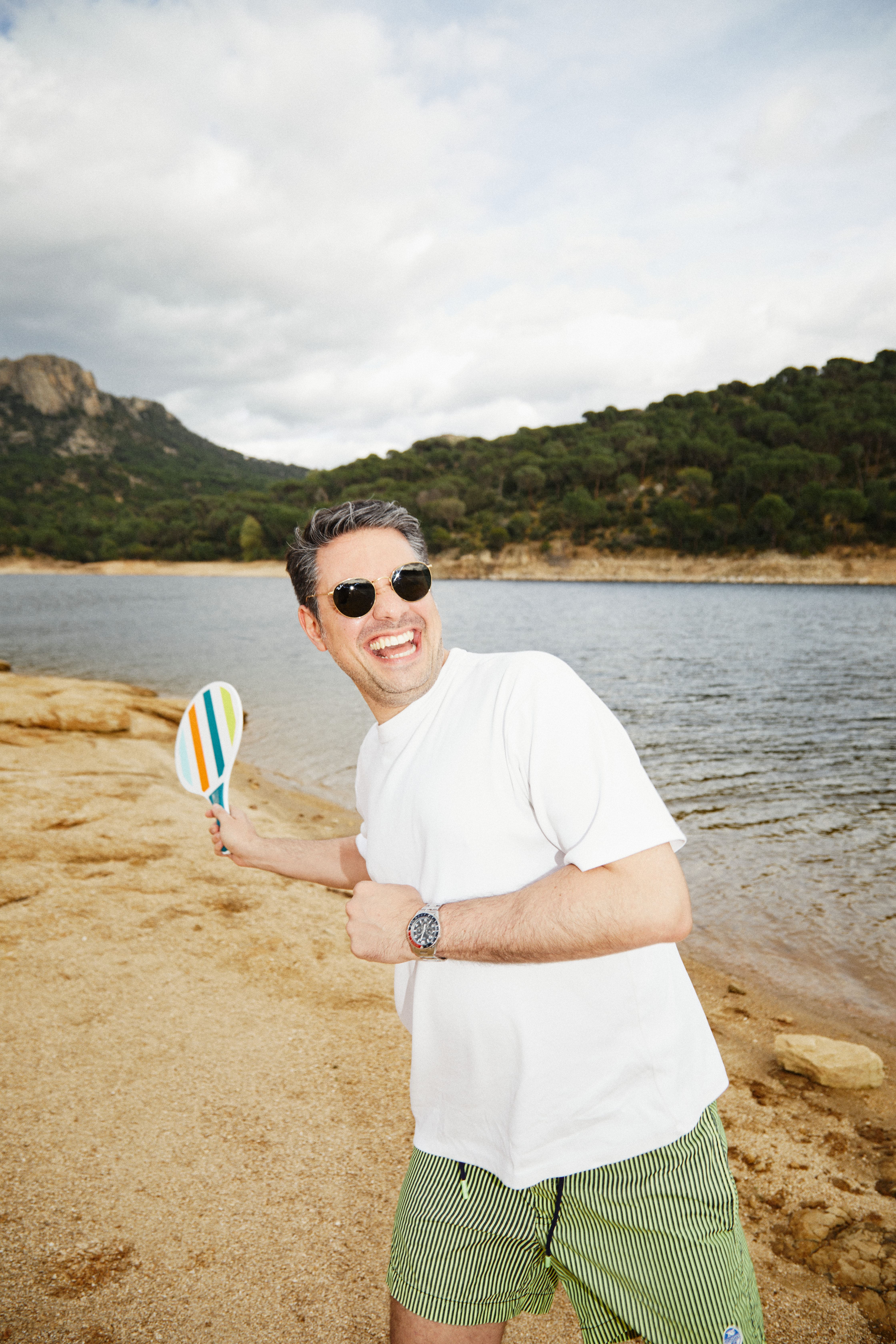 Smiling man wearing sunglasses and a white t-shirt, holding a colorful paddle on a sandy lakeshore with hills in the background.