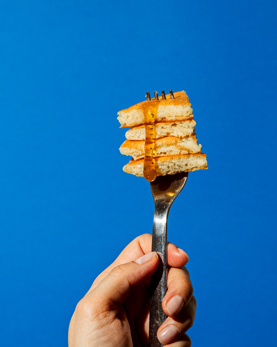 Hand holding a fork with four stacked small pancakes drizzled with honey against a blue background.