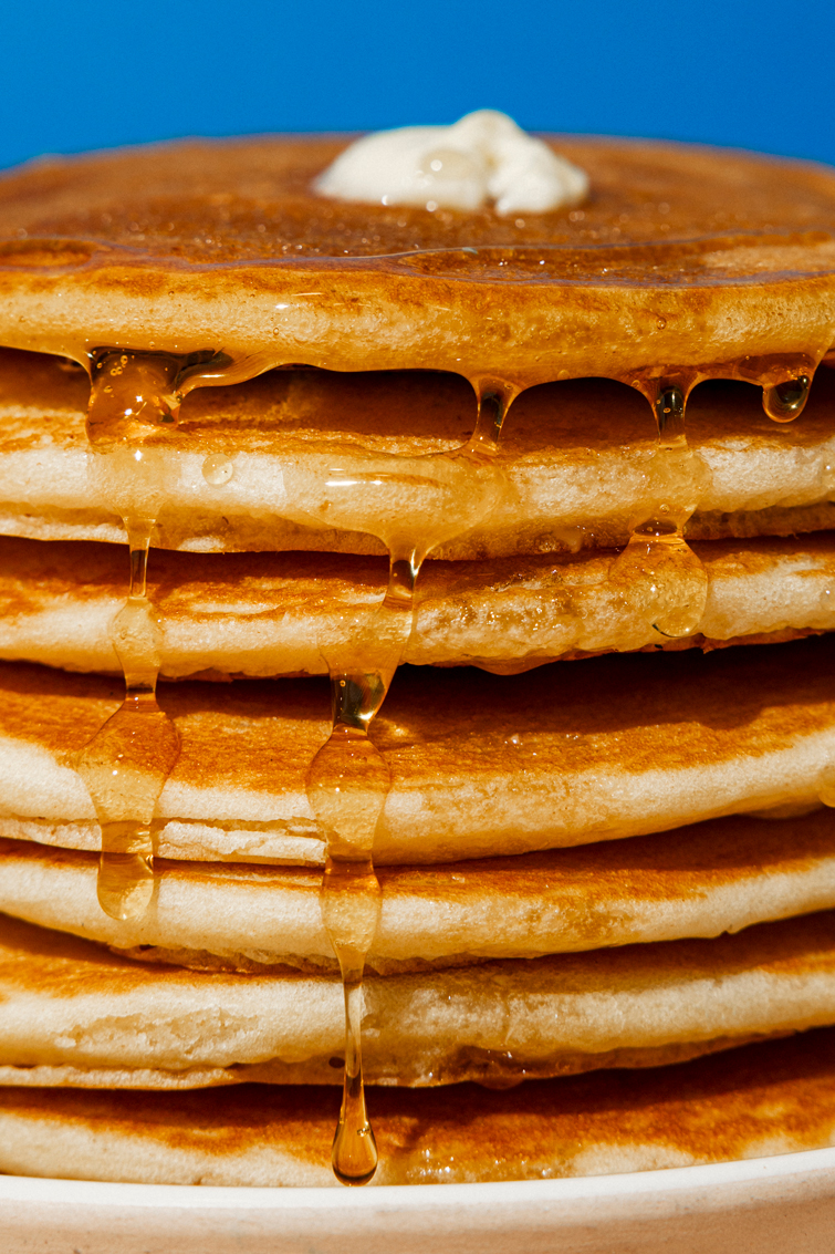 Close-up of a stack of golden pancakes with butter on top and syrup dripping down the sides.