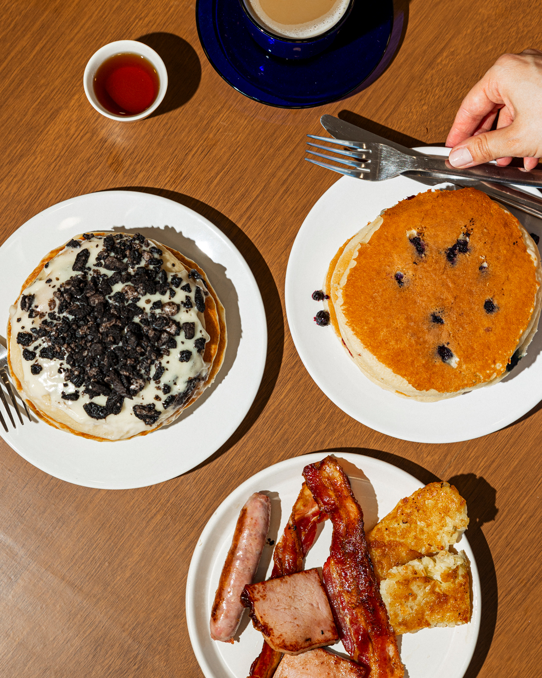 Top view of a breakfast table with two plates of pancakes, one with cream and cookie crumbles and the other with blueberries, alongside a plate with sausage, bacon, ham, and hash browns, a cup of coffee, and syrup on a wooden surface.