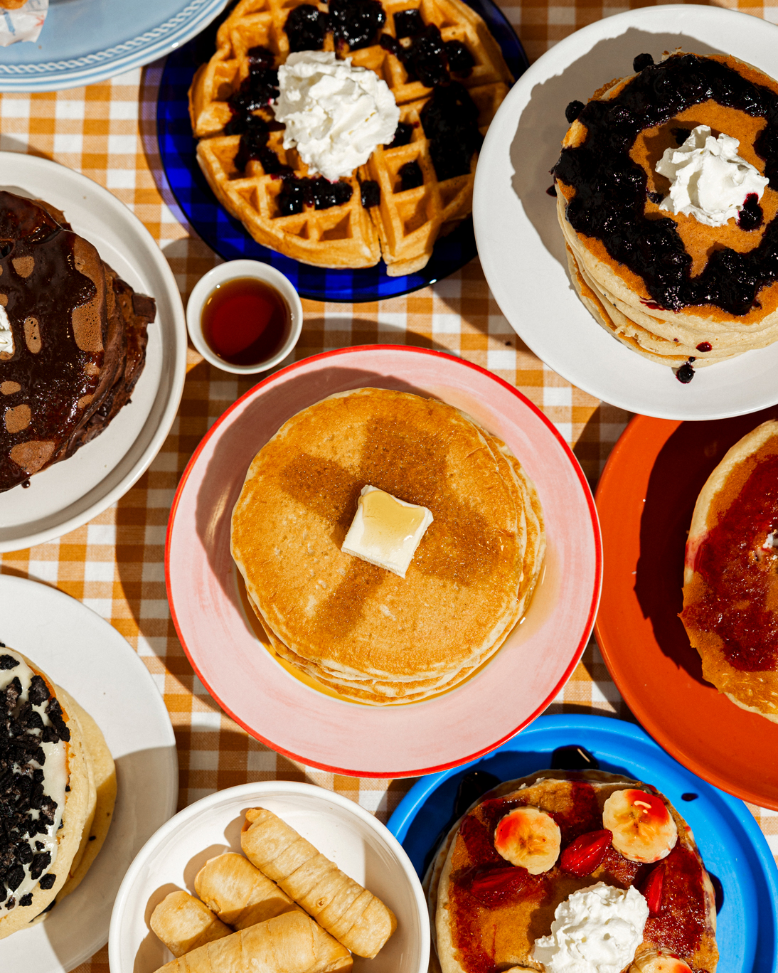 Overhead view of assorted pancakes and waffles with toppings including syrup, whipped cream, chocolate, blueberries, bananas, strawberries, and three bread rolls on a checkered tablecloth.