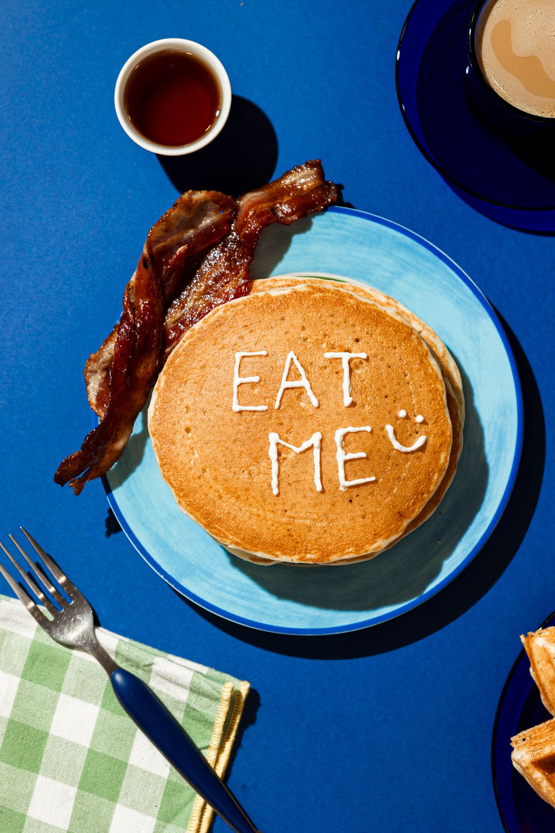 Stack of pancakes with 'EAT ME :)' written in syrup, served with two strips of bacon, syrup cup, and coffee on a blue table.