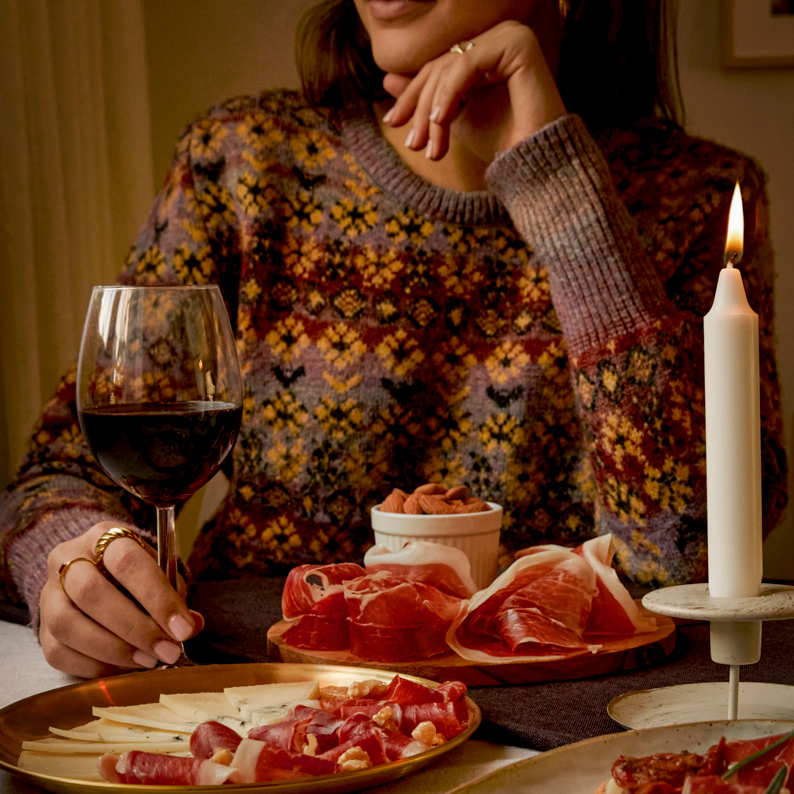 Person in a patterned sweater sitting at a table with a glass of red wine, plates of cured meats and cheese, a small bowl of nuts, and a lit white candle.