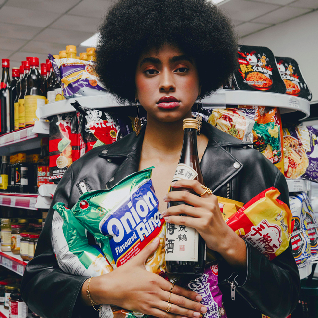Person with curly hair wearing a black leather jacket holding snacks and a bottle in a store aisle.