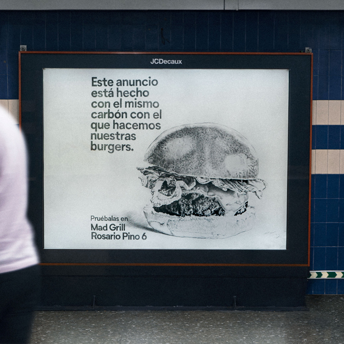 Black and white sketch of a burger with text in Spanish promoting Mad Grill restaurant, stating the ad is made with the same charcoal used for their burgers.