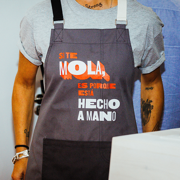 Person wearing a gray apron with text that reads 'SI TE MOLA, ES PORQUE ESTÁ HECHO A MANO' over a light gray shirt.